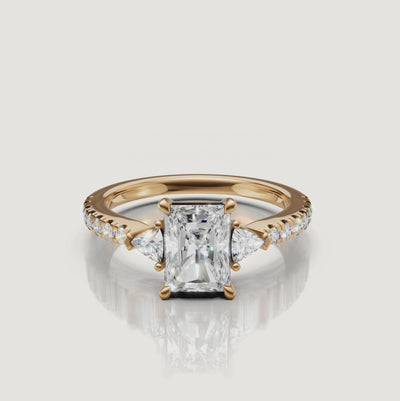 A 360-degree rotation of a Rual Diamonds radiant-cut three-stone ring in 14k yellow gold, showcasing the brilliant trillion side stones and shimmering pavé shank
