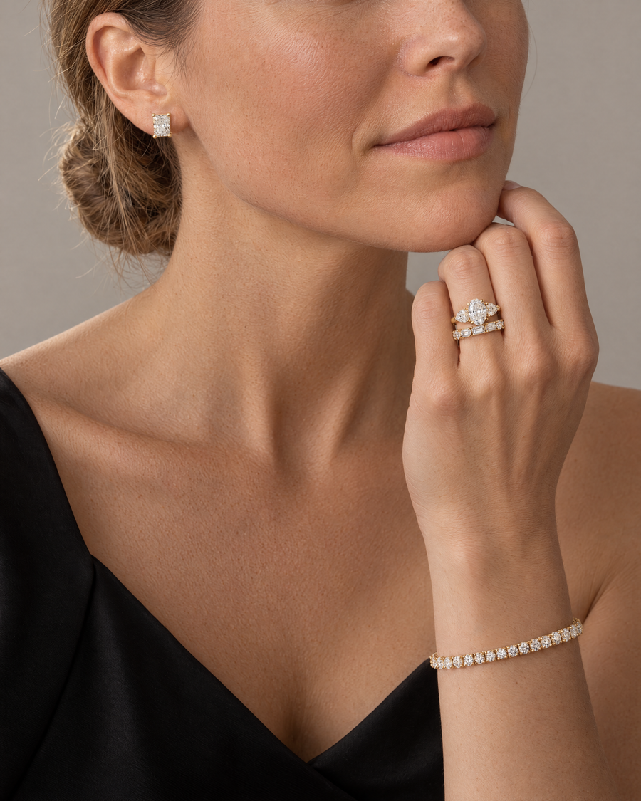 Woman wearing an oval three-stone lab-grown diamond engagement ring, a wedding band and radiant stud earrings in 14K yellow gold